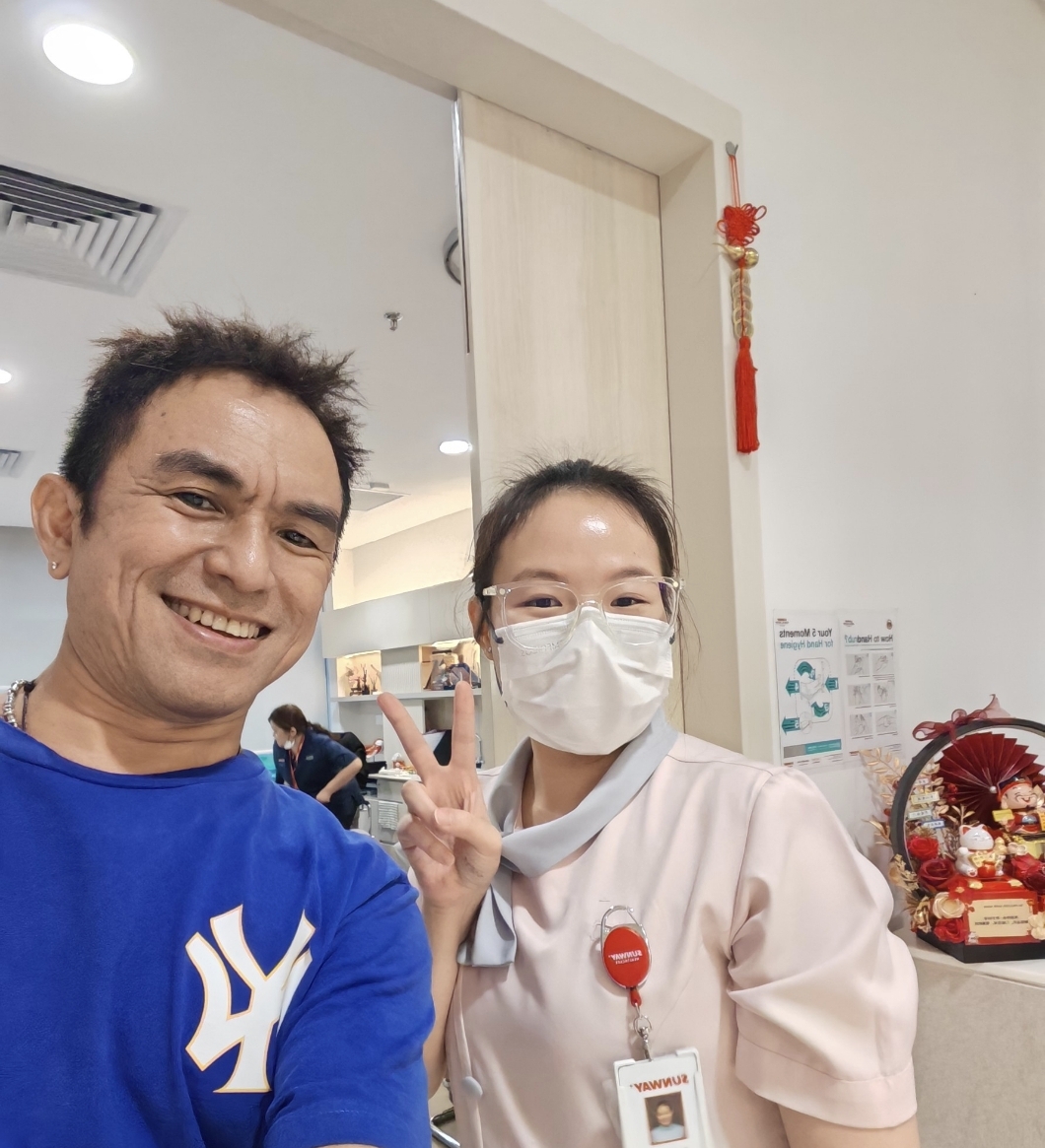 Two people take a selfie indoors: a smiling man in a blue NY shirt and a nurse in a beige uniform with a mask, making a peace sign.